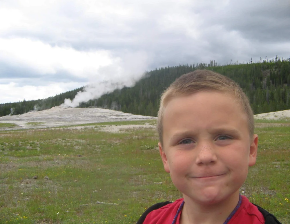 Boy standing in front of a geyser, meadow, and forest in the source photograph